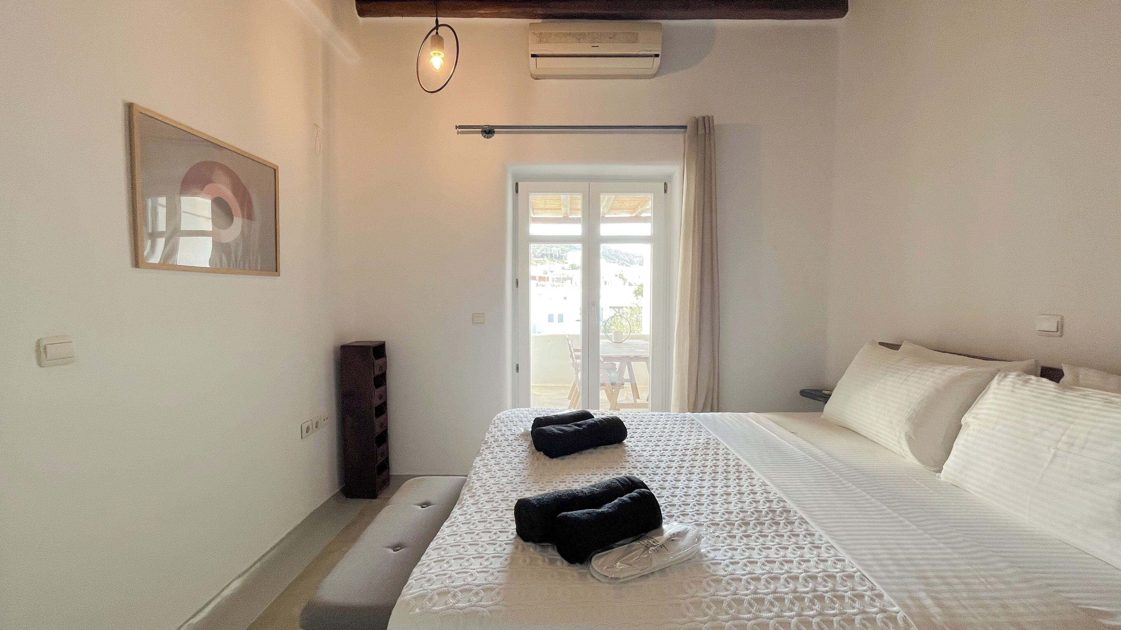 A bedroom with a bed, a bench, a wall-mounted air conditioner, a framed picture, and a door leading to a balcony.