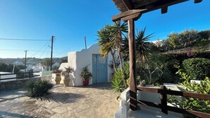 A white building with a blue door, surrounded by potted plants and a stone patio. - Pool House in Ornos (Mykonos)