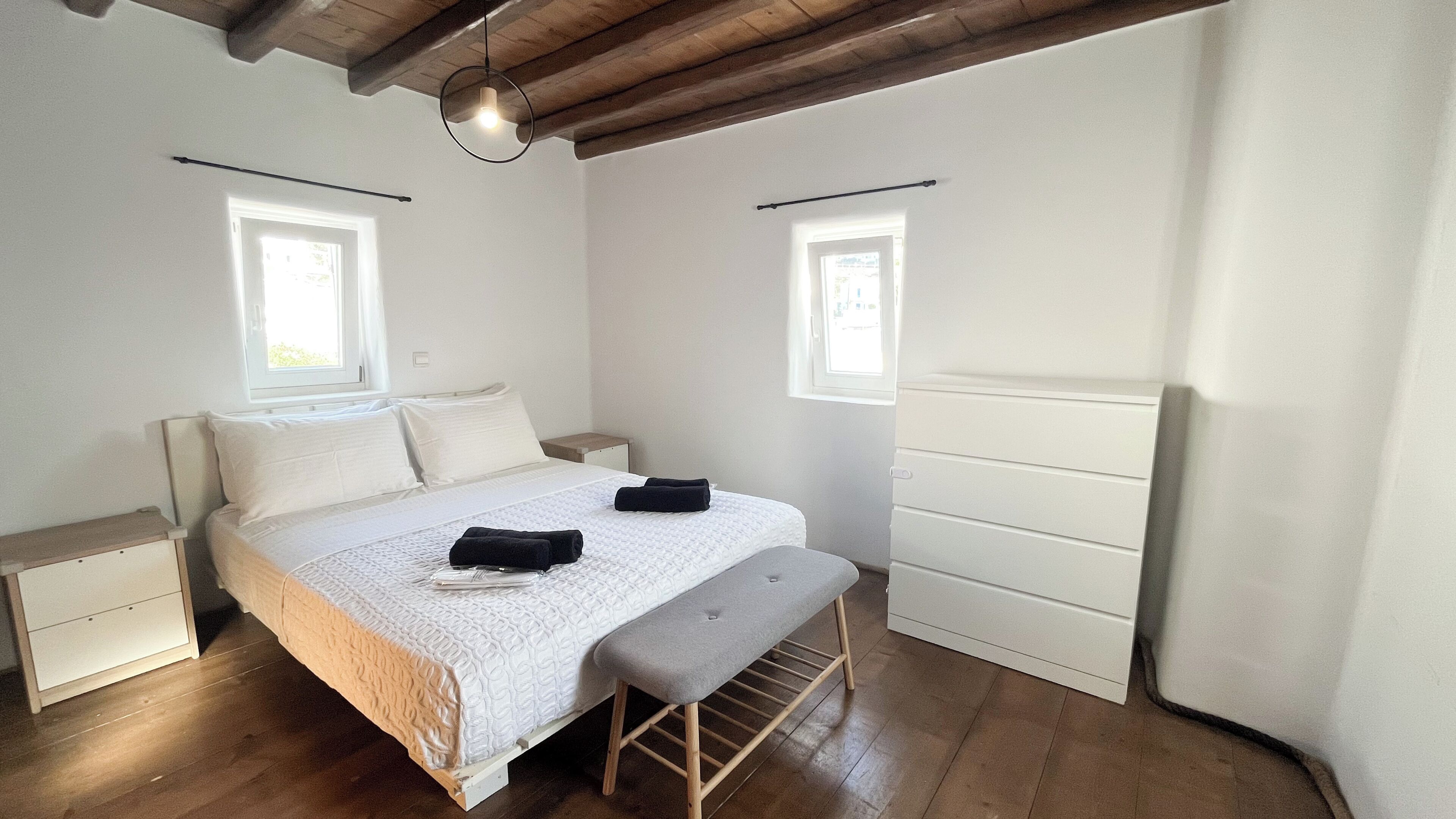 A bedroom with a bed, bench, bedside tables, and two windows.