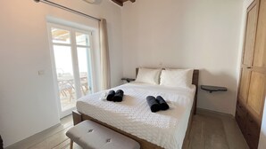 A bedroom with a bed, bench, bedside table, and a window with curtains. - Pool House in Ornos (Mykonos)