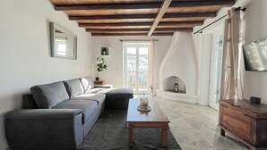 A living room with a grey sofa, a wooden coffee table, a stone fireplace, and a television mounted on the wall. - Pool House in Ornos (Mykonos)