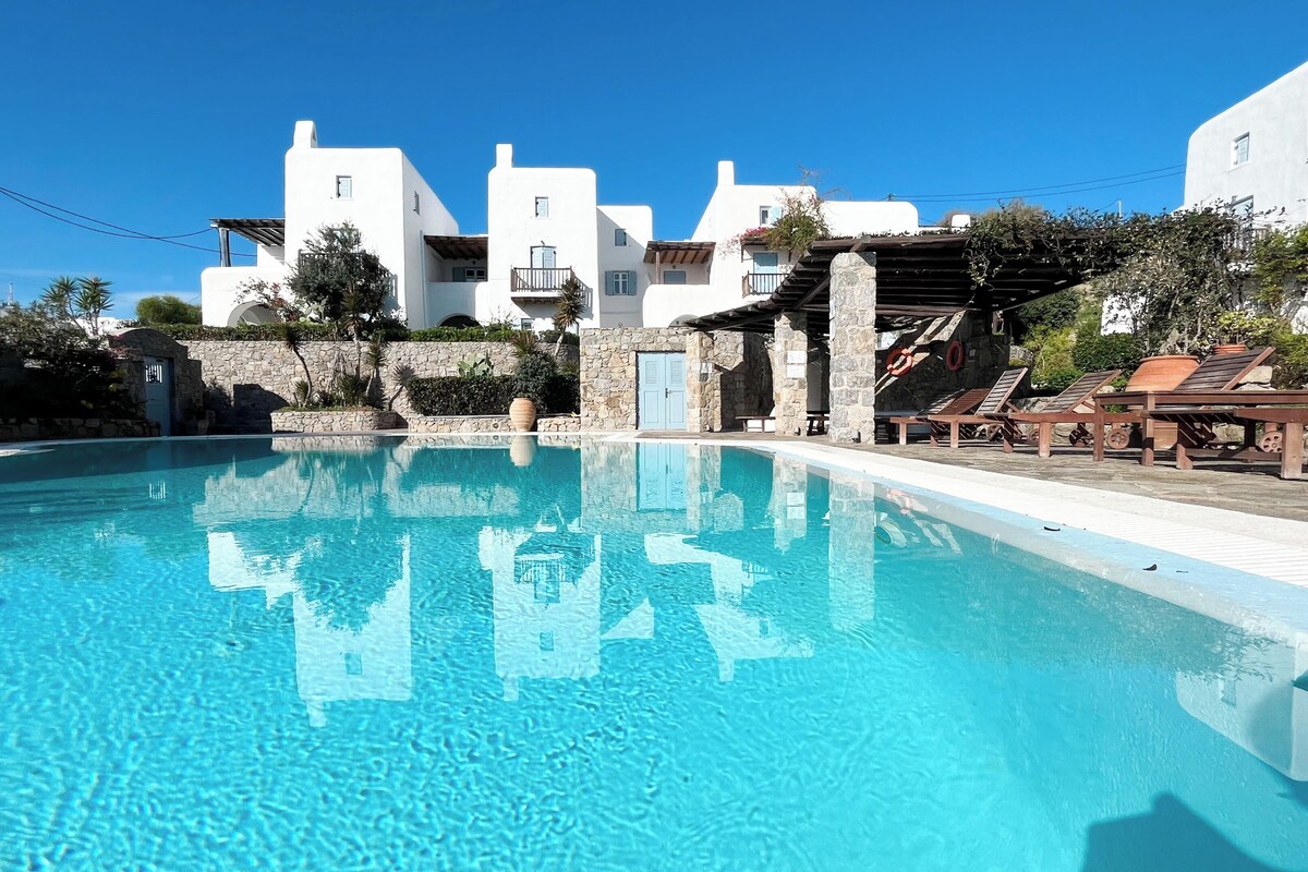 A clear swimming pool with a stone wall and a wooden deck with lounge chairs.