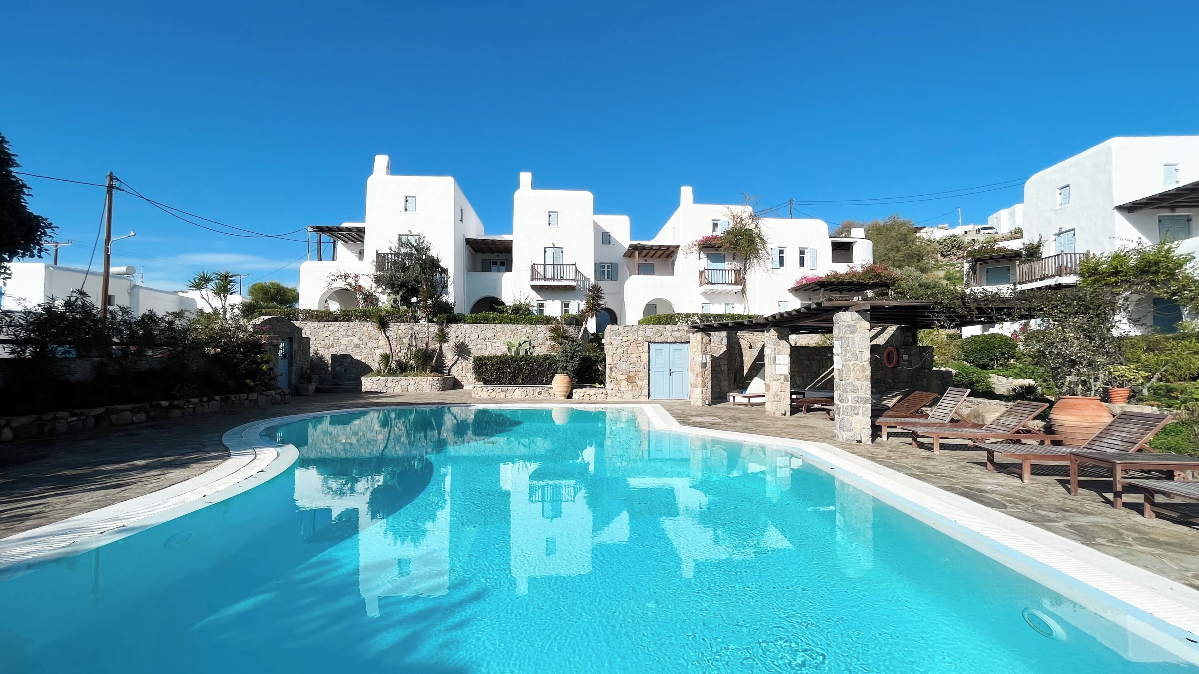A swimming pool with a stone wall and lounge chairs, surrounded by white buildings with balconies.
