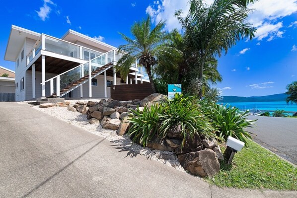 A modern two-story house with a staircase leading to a balcony, surrounded by rocks and tropical plants.