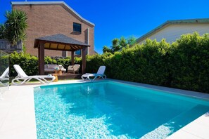 A swimming pool with lounge chairs, a covered seating area, and a brick building in the background.