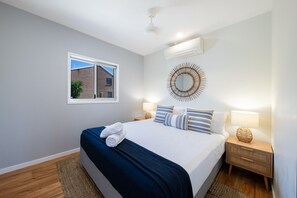 A bedroom with a bed, a window, a wall-mounted mirror, and a bedside table with a lamp.