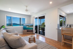 A modern living room with a sofa, a coffee table, and a TV. There is a balcony with a view of the sea.