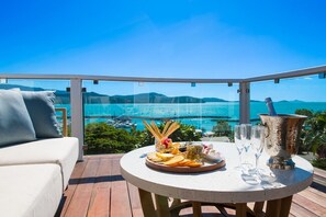 Een ronde tafel met diverse hapjes en drankjes, een champagnefles en een glas op een balkon met uitzicht op een wateroppervlak en boten.