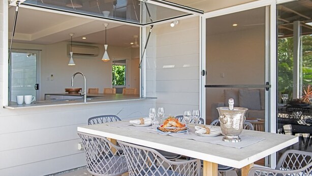 An outdoor dining area with a table set for two, surrounded by chairs, and a large window overlooking a garden. - Nautical Views - Airlie Beach (Airlie Beach)