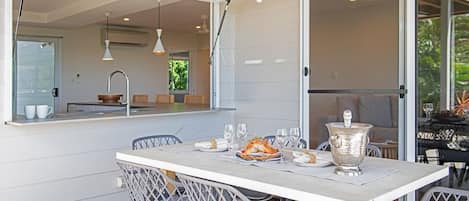 An outdoor dining area with a table set for two, surrounded by chairs, and a large window overlooking a garden.
