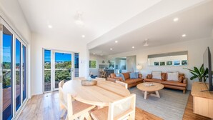 A spacious living area with a wooden dining table, sofas, and a coffee table. There is a sliding glass door leading to an outdoor area. - Nautical Views - Airlie Beach (Airlie Beach)