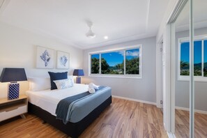 Een slaapkamer met een groot bed, twee nachtkastjes, een plafondventilator en een raam met uitzicht op groen.