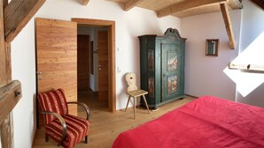 A room with a wooden ceiling, a red bed, a wooden chair, a green cabinet with artwork, and a door leading to another room. - Stylish vacation apartment in an old farmhouse - south view (Kirchbach)