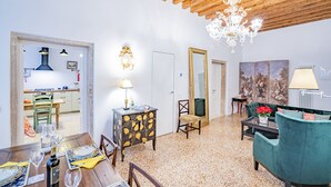 Zona pranzo con tavolo in legno, sedie e lampadario. Si intravede una cucina attraverso una porta aperta.
