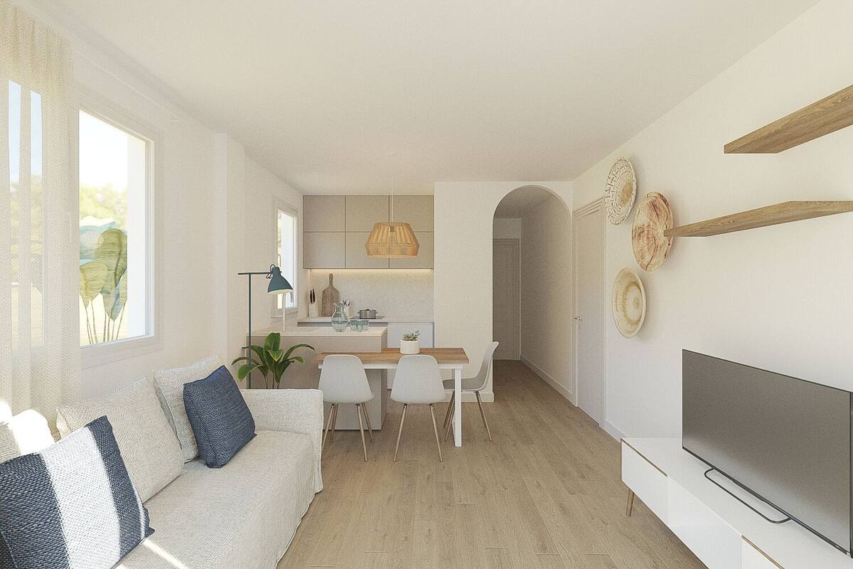 A modern living room with a sofa, dining table, and chairs. There is a kitchen area with cabinets and a hanging light fixture. A television is mounted on the wall.