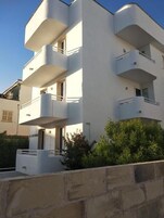 A modern multi-story building with balconies, a stone wall in the foreground, and a clear sky.