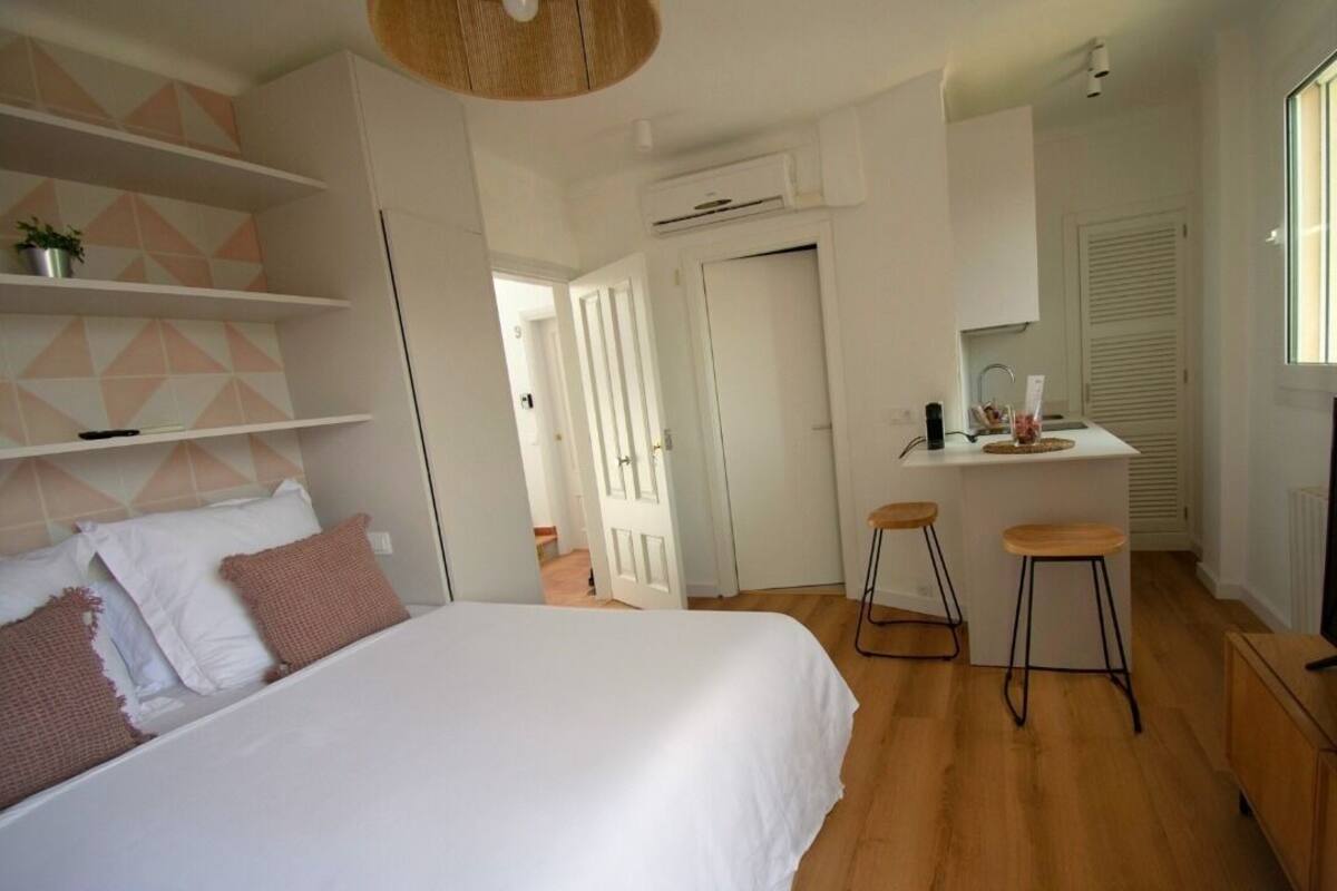A modern bedroom with a large bed, a small kitchenette, and a bathroom visible in the background.