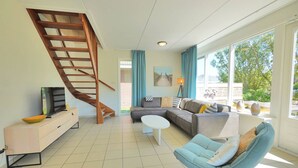 En moderne stue med en trætrappe, en modulsofa, en blå lænestol og et stort vindue med udsigt til grønne områder.