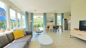 En moderne stue med sofa, sofabord, spiseområde med stole, fjernsyn og et køkken med køleskab.