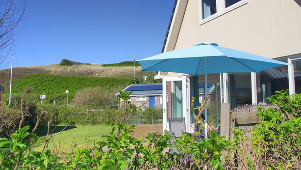 Et moderne hus med en blå parasol, græsplæne og en bakke i baggrunden.