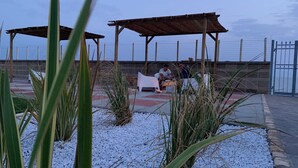 Een overdekt terras met tafels en stoelen, omringd door witte kiezelstenen en planten.