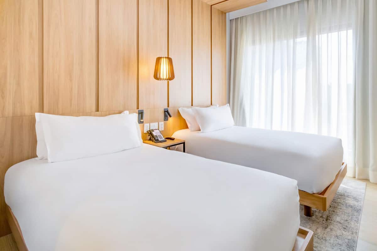 Une chambre d’hôtel avec deux lits, un mur lambrissé en bois, une lampe de chevet et une fenêtre avec des rideaux.
