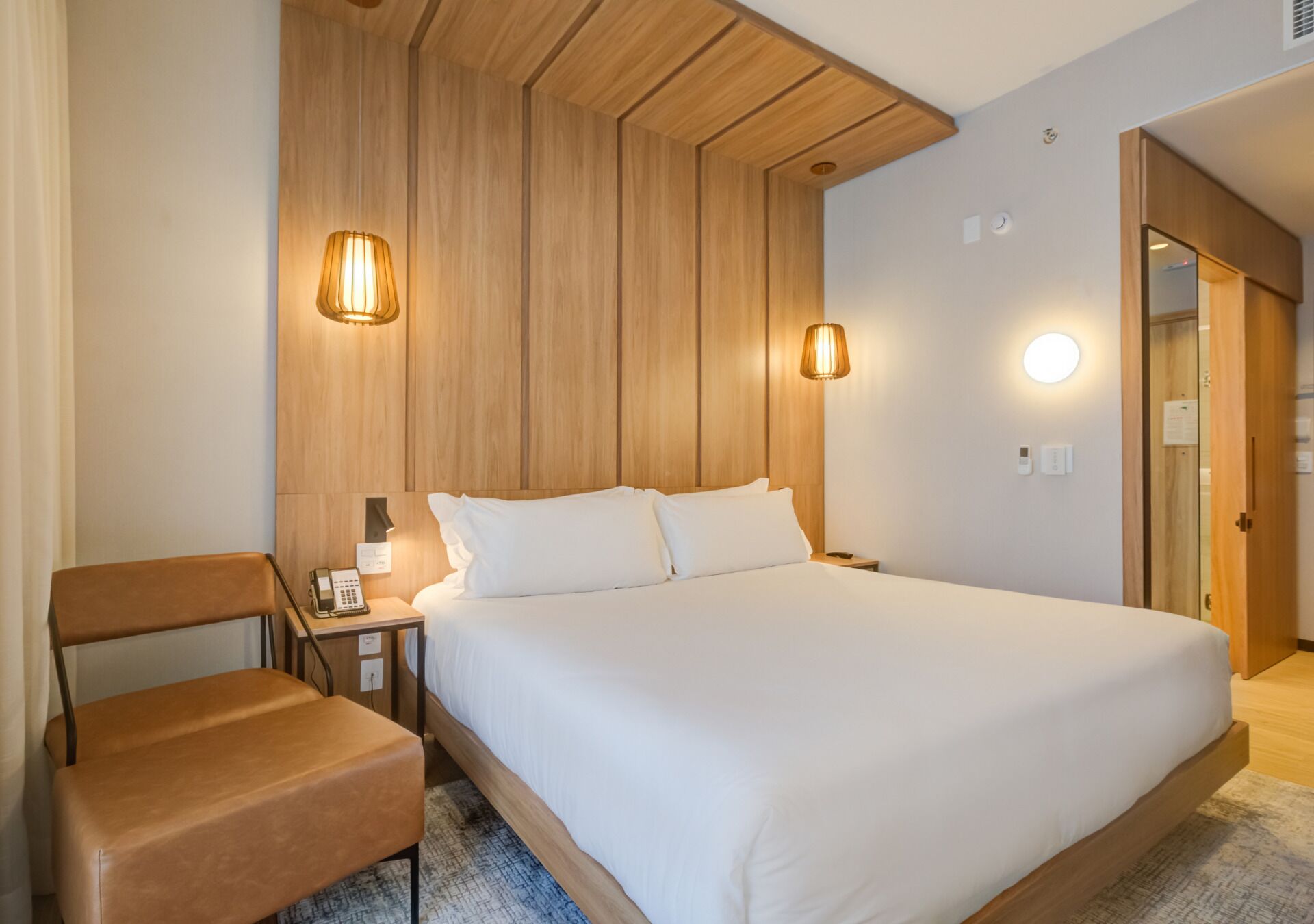 A hotel room with a large bed, a wooden headboard, two bedside lamps, a nightstand with a phone, and a brown leather chair.