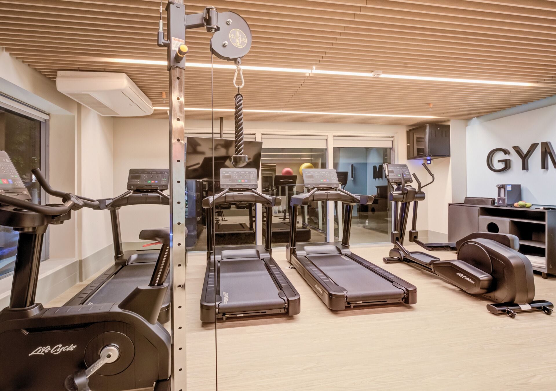 Un gimnasio con cintas de correr, una máquina de pesas y un equipo de aire acondicionado instalado en la pared.