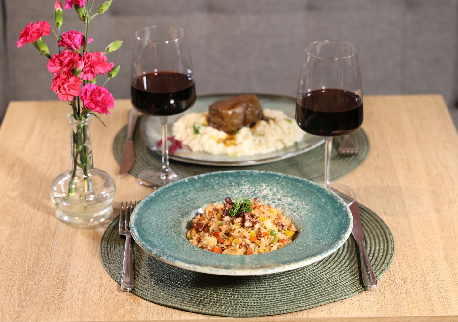 Ein Tisch mit einem Teller Essen, zwei Gläsern Rotwein und einer Vase mit rosa Blumen.