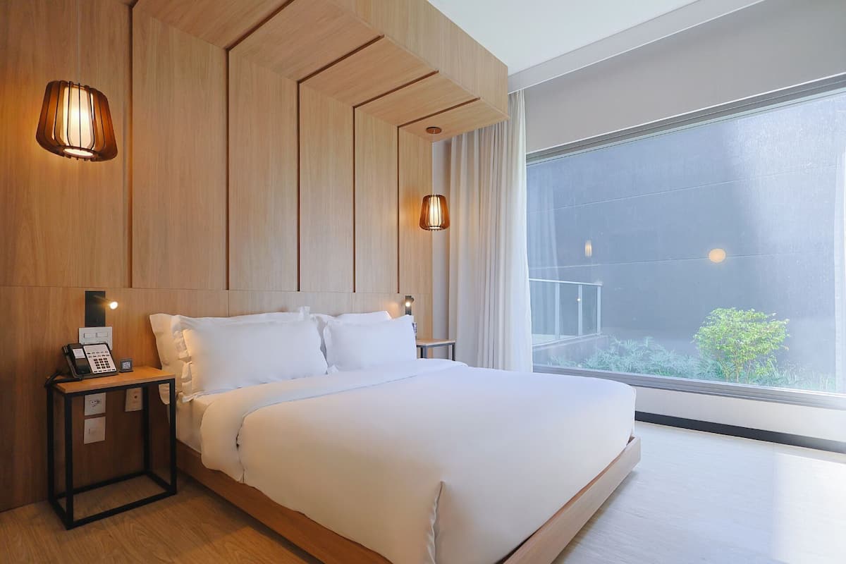 Une chambre d’hôtel moderne avec un grand lit, des murs recouverts de panneaux de bois, une table de chevet avec un téléphone et une grande fenêtre donnant sur un paysage verdoyant.