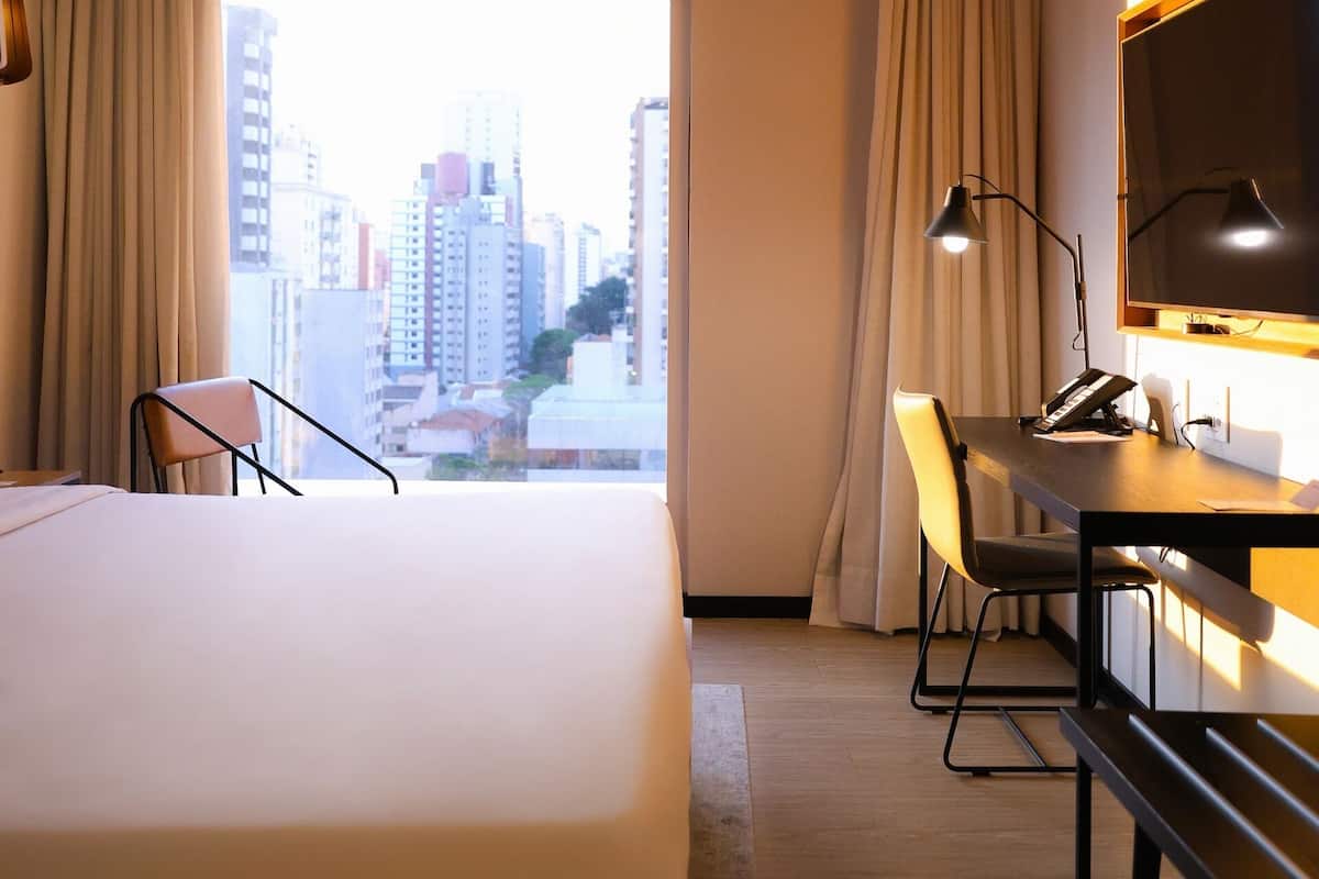 Une chambre d’hôtel avec un lit, un bureau doté d’une lampe, une chaise et une vue sur les bâtiments de la ville.