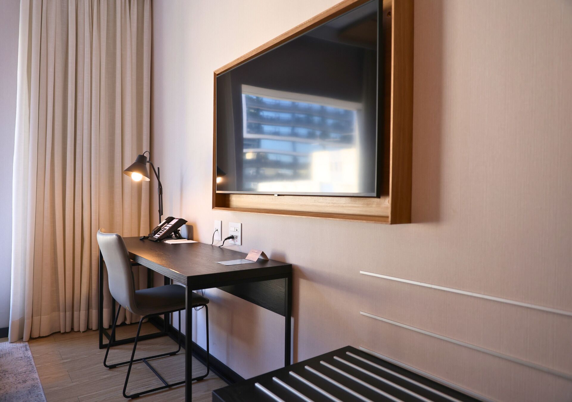 Ett hotellrum med ett skrivbord, en stol, en lampa och ett stort fönster med utsikt över byggnader.