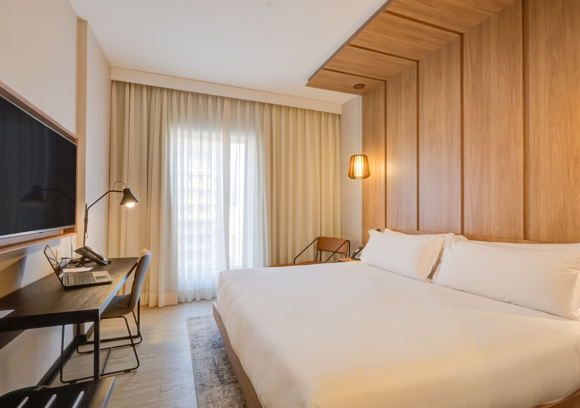 A hotel room with a bed, a desk with a laptop, a TV, a lamp, and a window with curtains.