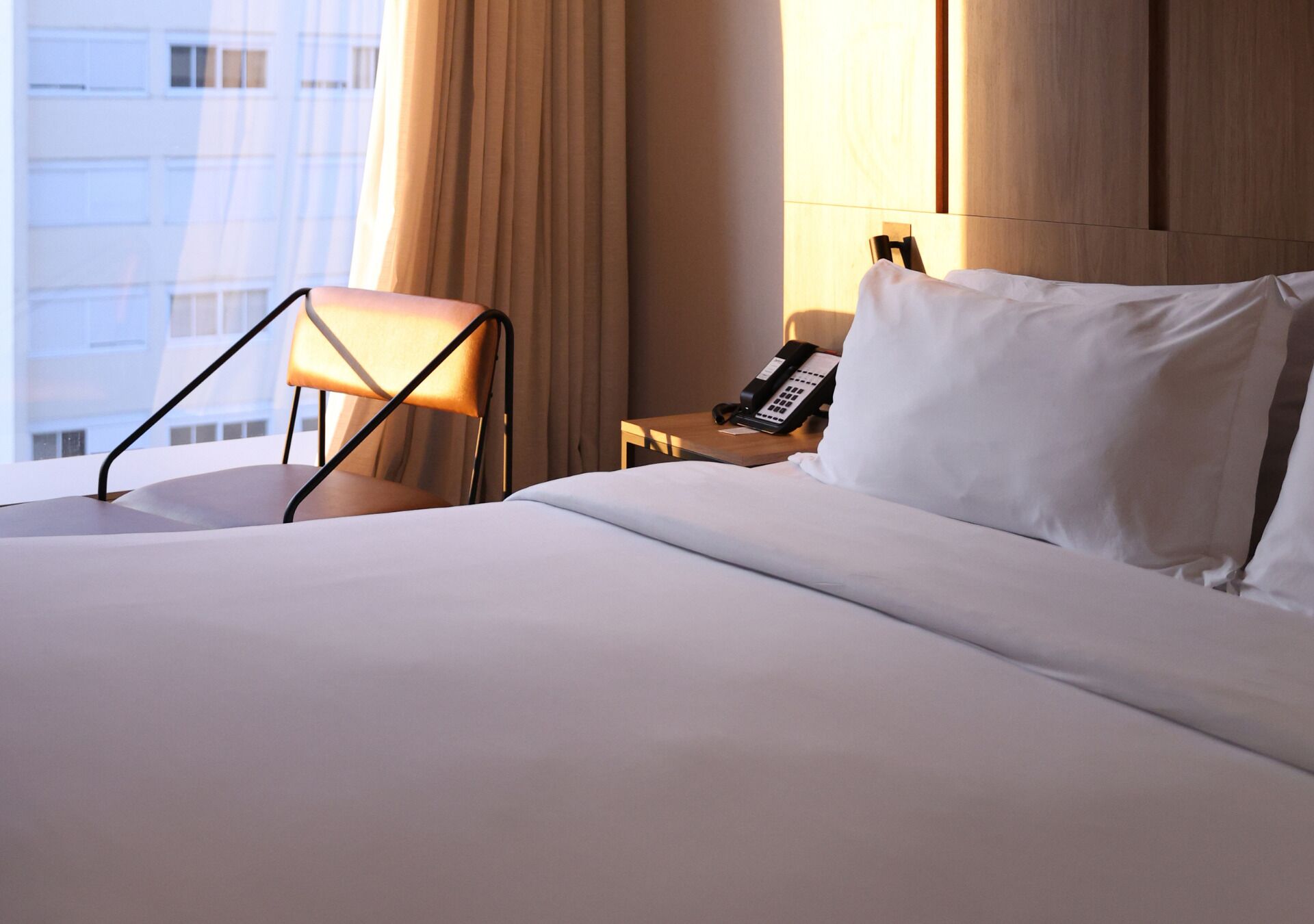 A hotel room with a bed, a chair, a bedside table with a phone, and a window with curtains.