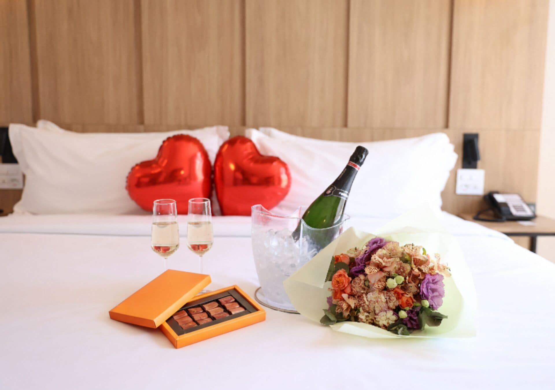 A bed with a bouquet of flowers, two glasses of champagne, a bottle of champagne, and a box of chocolates.
