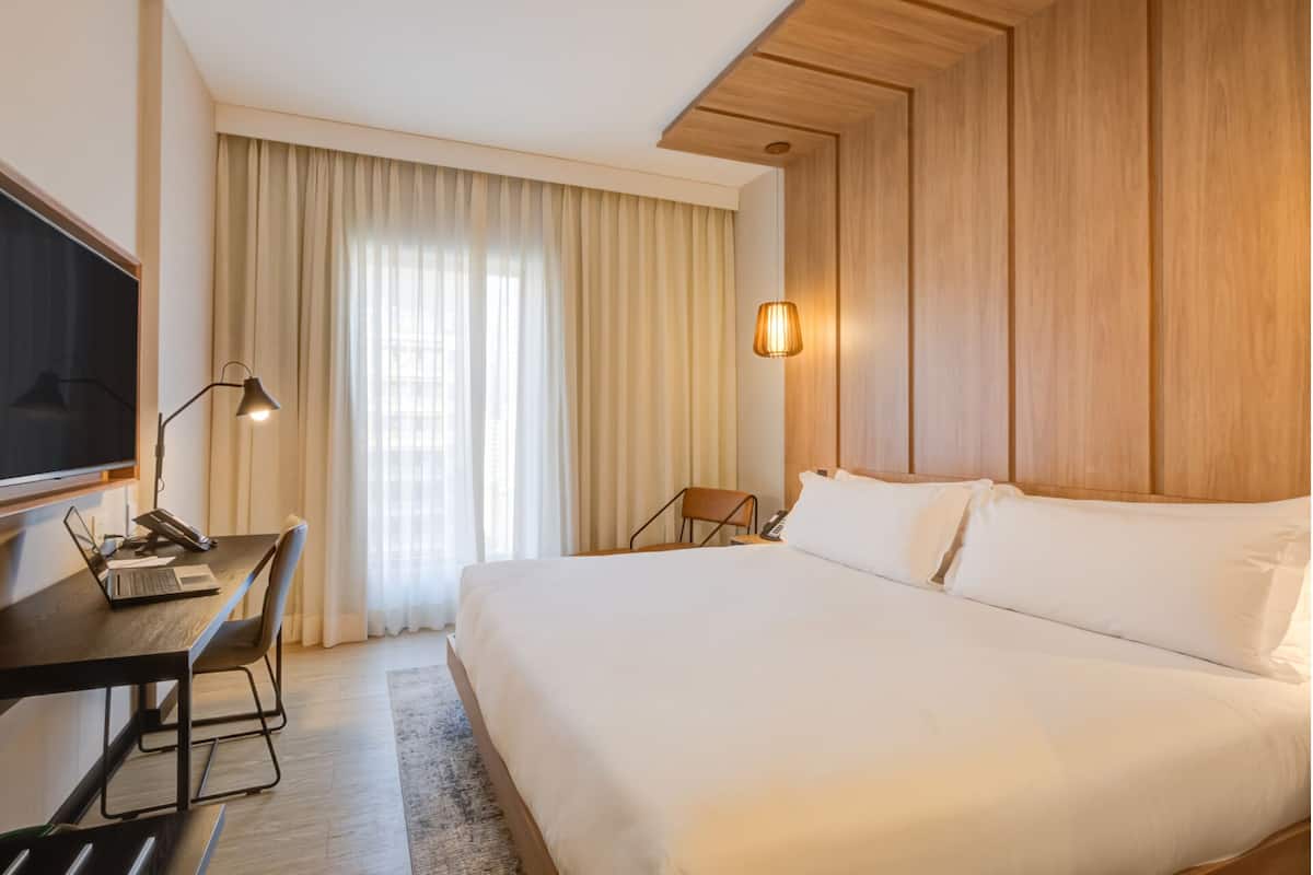 Une chambre d’hôtel avec un grand lit, un bureau avec un ordinateur portable, une chaise, une télévision et une fenêtre avec des rideaux.