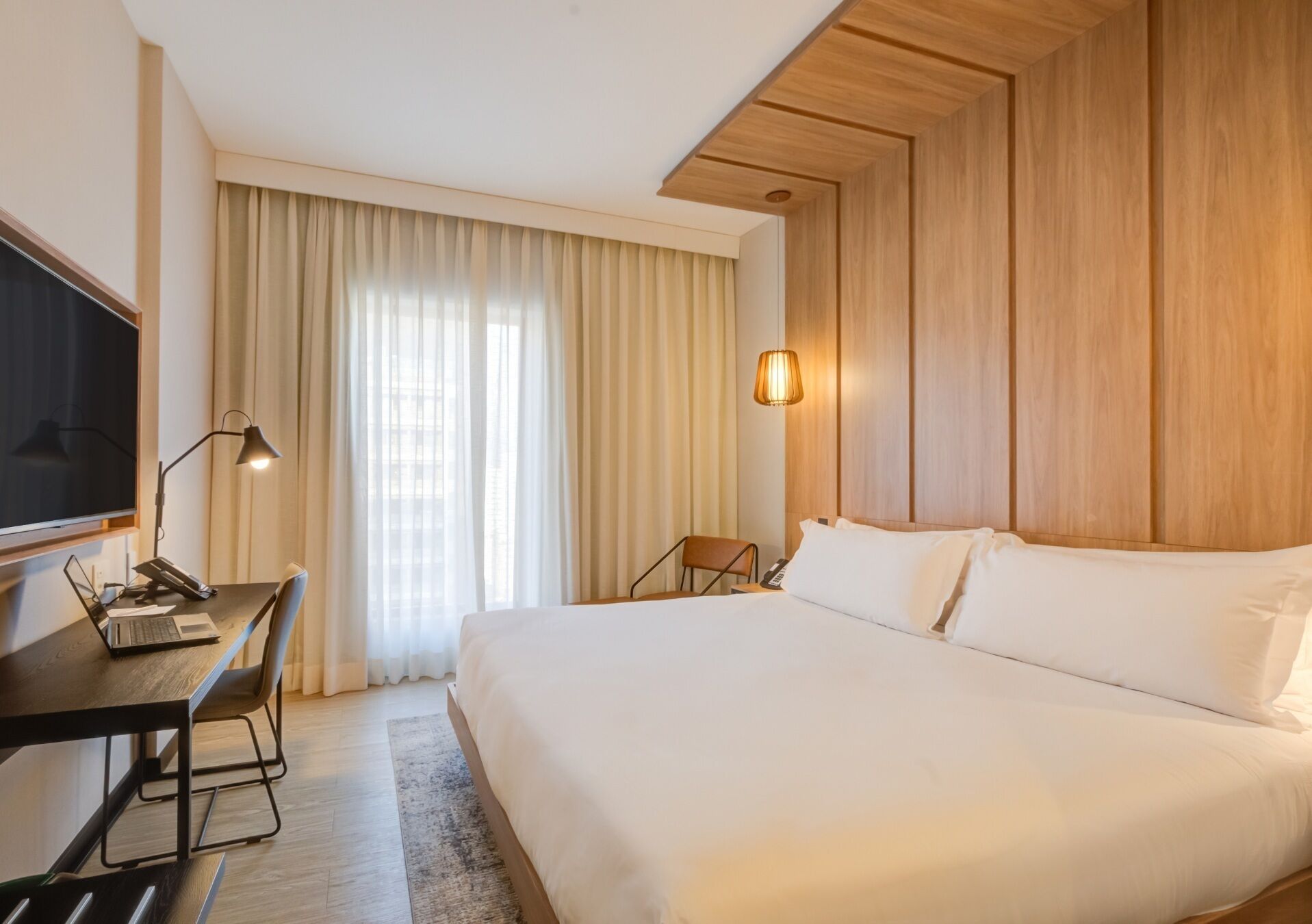 A hotel room with a large bed, a desk with a laptop, a chair, a TV, and a window with curtains.