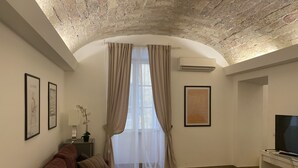 Ein Wohnzimmer mit einem braunen Sofa, einem Couchtisch mit Pflanze, einem Fernseher auf einem Schrank und einem großen Fenster mit Vorhängen.