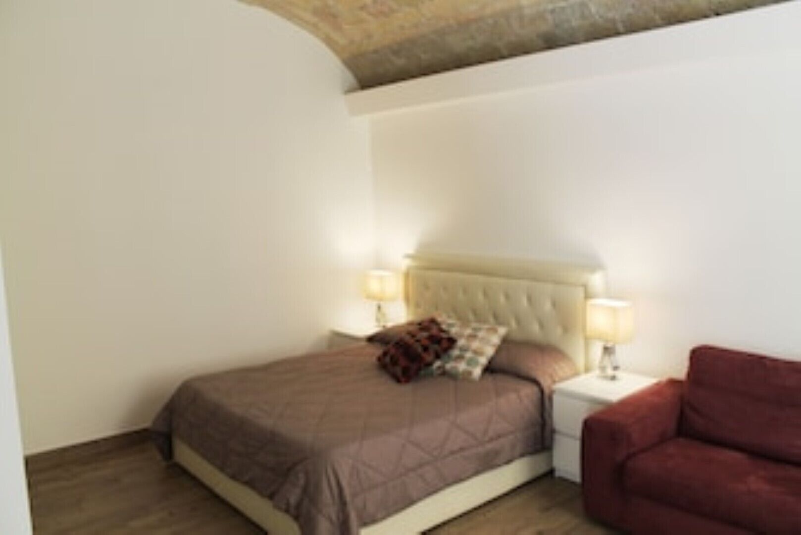 A bedroom with a bed, two bedside lamps, a red chair, and a wooden floor.