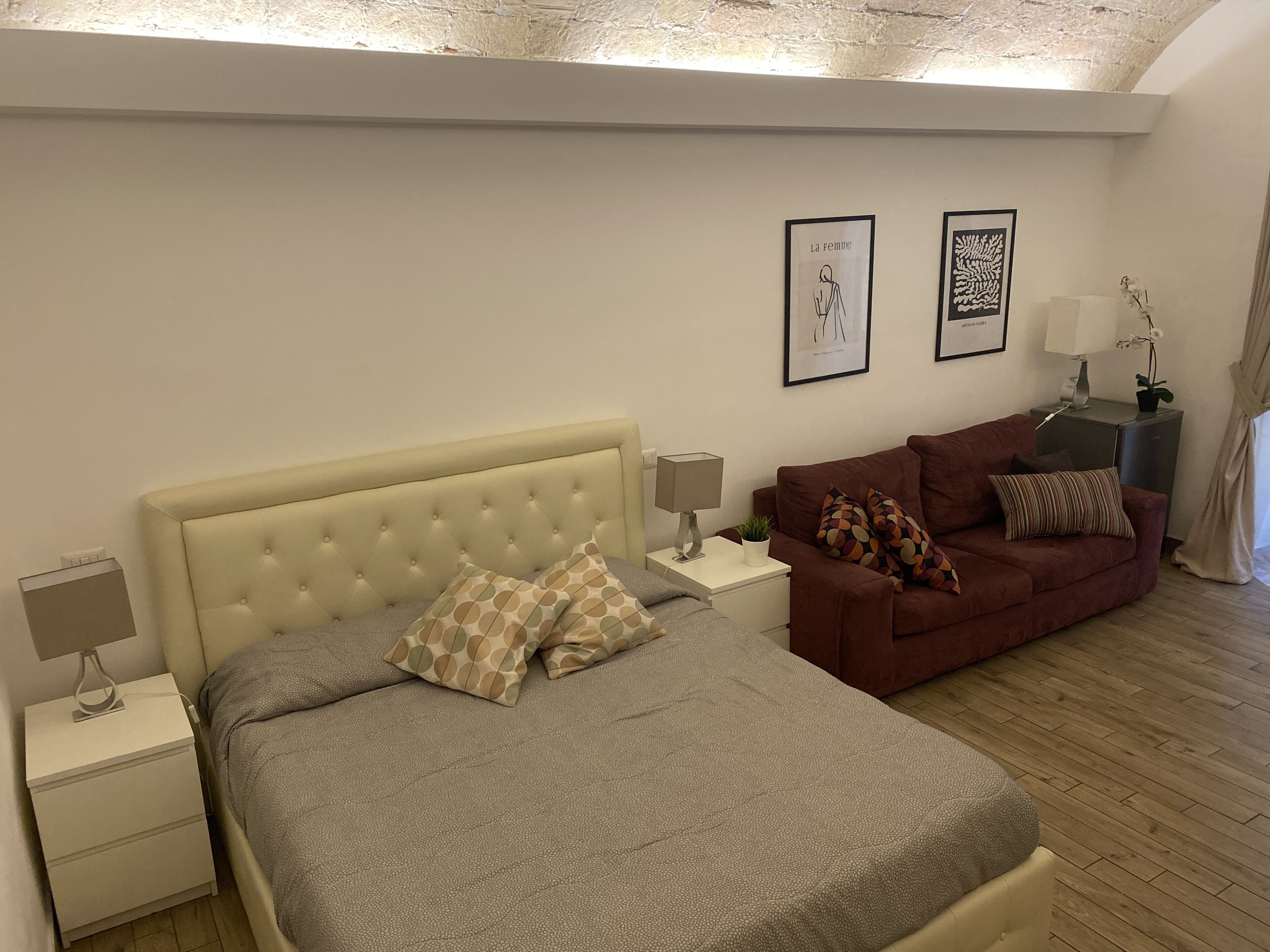 A bedroom with a bed, a red sofa, a nightstand, a lamp, and two framed pictures on the wall.
