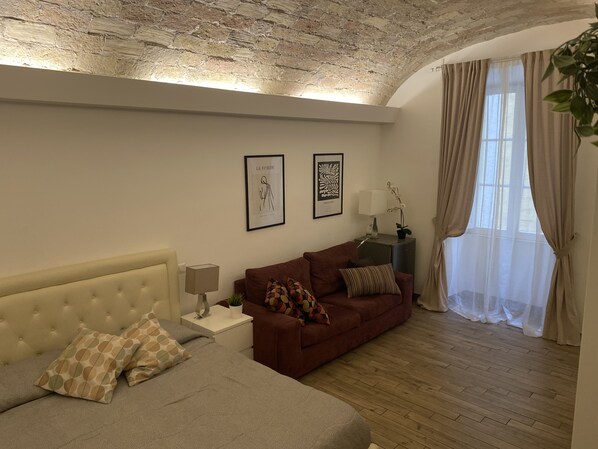 Ein modernes Wohnzimmer mit einem beigen Sofa, einem braunen Schlafsofa und einem Bett mit gemusterten Kissen. An der Wand hängen zwei gerahmte Kunstwerke und auf einem Beistelltisch steht eine Lampe.