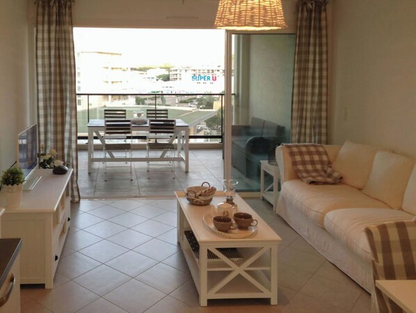 A living room with a sofa, a coffee table with cups, a TV, and a balcony with chairs.