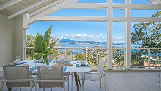 A dining area with a table set for two, overlooking a scenic view of a coastal town and mountains. - The 5 Tangarane apartments are located in Les Trois Ilets, overlooking the magni (Les Trois-Îlets)