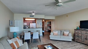 A living room with a sofa, a coffee table, and a TV. There is a dining area with a table and chairs. The room has a ceiling fan and a kitchen in the background. - Aqua 404 | Amazing Views, Incredible Amenities & Location! (Panama City Beach)
