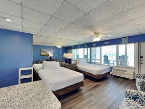 Une chambre d’hôtel avec deux lits, un ventilateur de plafond, une fenêtre avec des stores, un radiateur et une petite table avec une lampe.