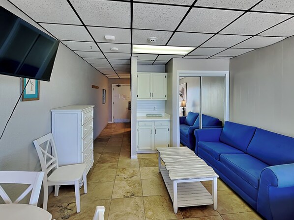 A living room with a blue sofa, white furniture, a flat-screen TV, and a kitchen area in the background.