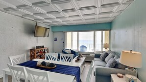 Una habitación con una mesa de comedor, sillas, un sofá, un televisor montado en la pared y una ventana con vistas.