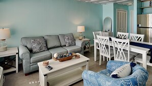 Una sala de estar con un sofá gris, sillas blancas, una mesita de centro y un sillón azul. Se observa una mesa de comedor con sillas, una lámpara y un espejo.