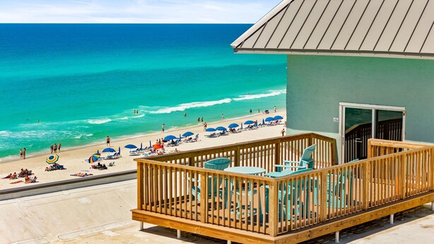 Una terraza de madera con sillas y mesas, con vista a una playa con sombrillas y gente.