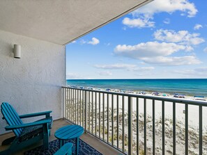 A balcony with a view of the beach, a chair, a small table, and a railing.